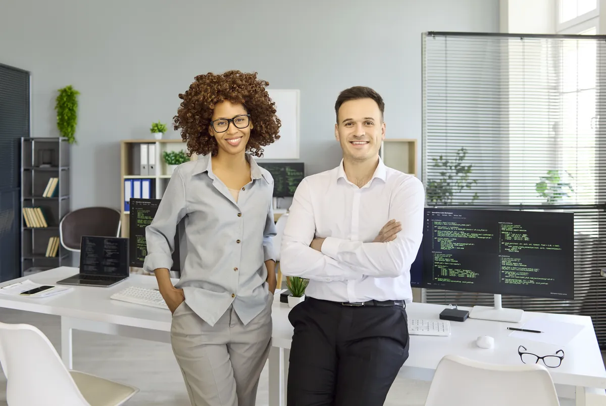 Two smiling IT professionals beside coding screens in a bright office