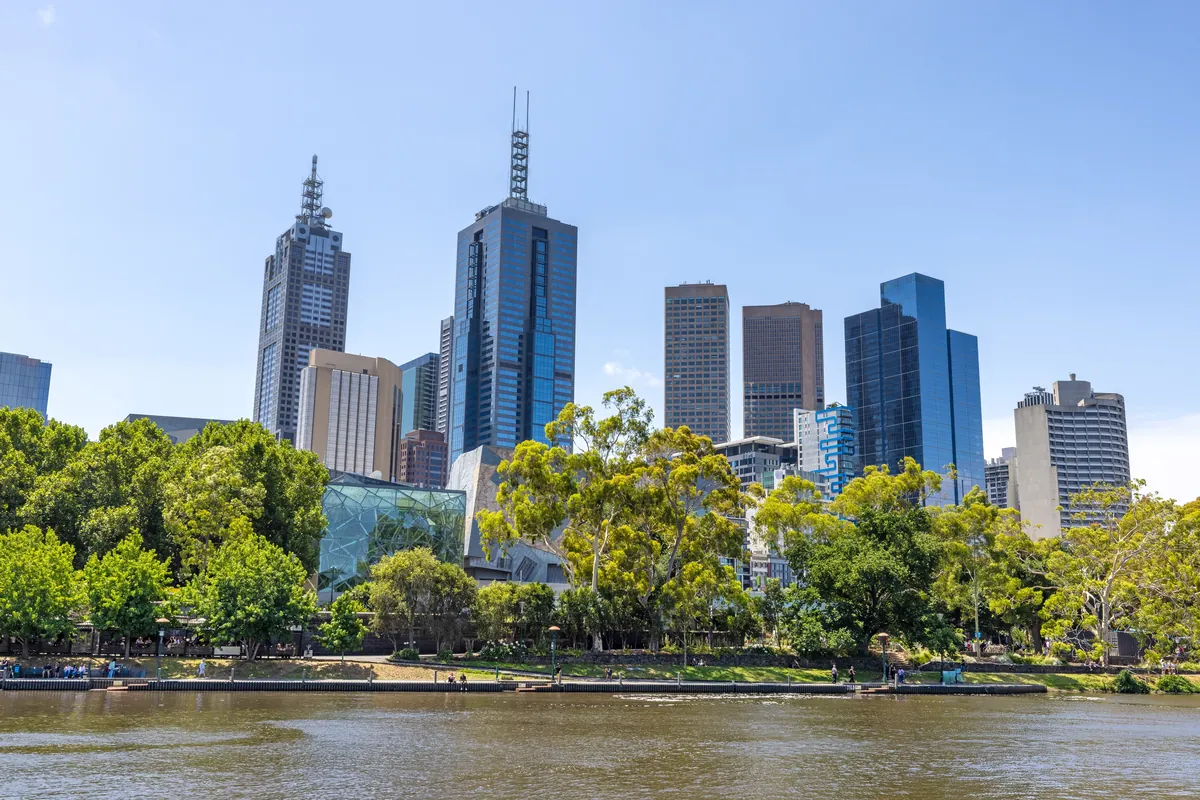 Melbourne city skyline above the Yarra River and green riverbank trees on a sunny day