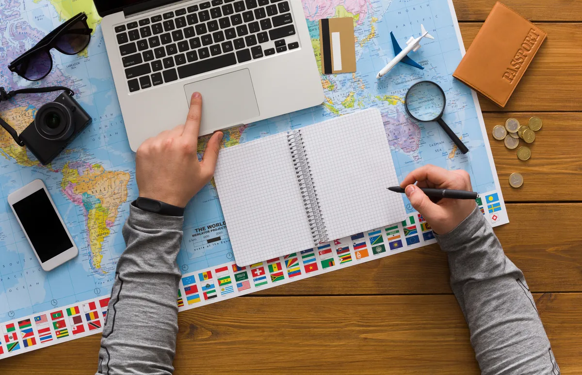 Hands writing in a notebook beside a laptop on a world map